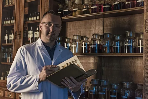 Man in lab coat reading a book in front of shelves of jars