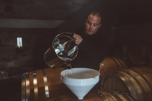 Distiller pouring from a glass jug into a funnel on a barrel