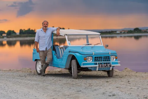 Man leaning on a vintage blue beach car at a Camargue salt lagoon at sunset
