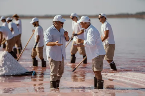 Salt workers in white raking sea salt in a pink Camargue salt pond