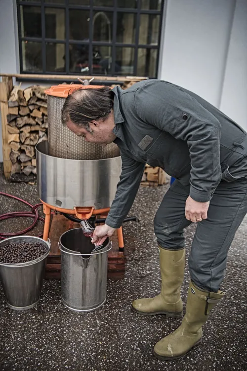 Man bending over a stainless steel vat next to a stack of firewood