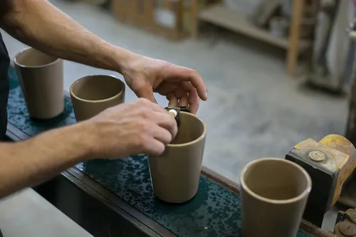 Hands finishing unfired stoneware cups on a workbench