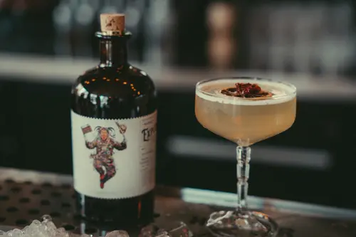 Dresdner Allerlei cocktail in a coupe next to an illustrated gin bottle